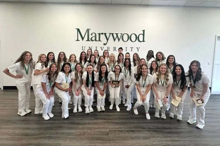 A group of nursing students in their white uniforms, standing in front of a white wall with a green Marywood wordmark on it.