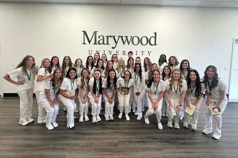 A group of nursing students in their white uniforms, standing in front of a white wall with a green Marywood wordmark on it.