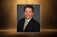 A formal headshot of a man in a suit against a gold glitter background