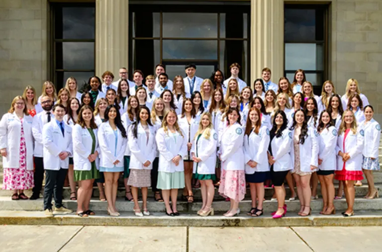 A group of physician assistant students gather in white coats in front of a building with stone columns.