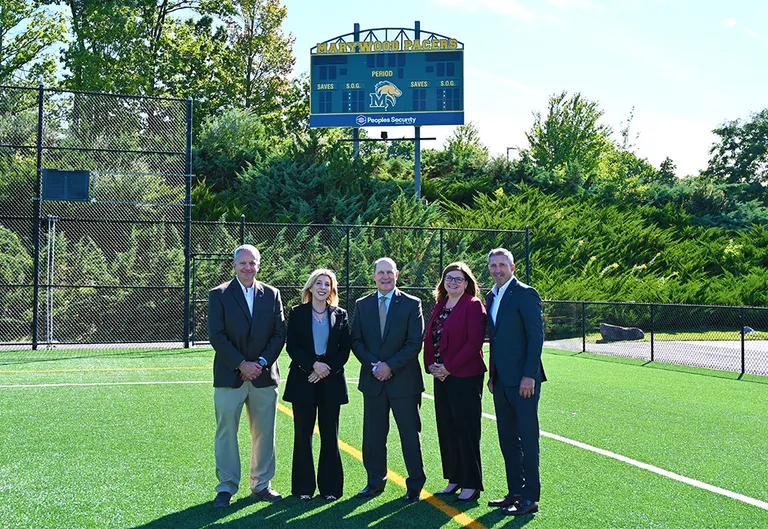five people standing on a green athletic field in front of a blue scoreboard