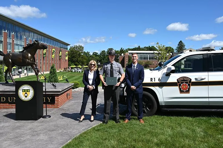 A woman in a navy suit and a man in a navy suit with a state trooper in the middle near a bronze horse statue, a state police SUV, and blue sky background.