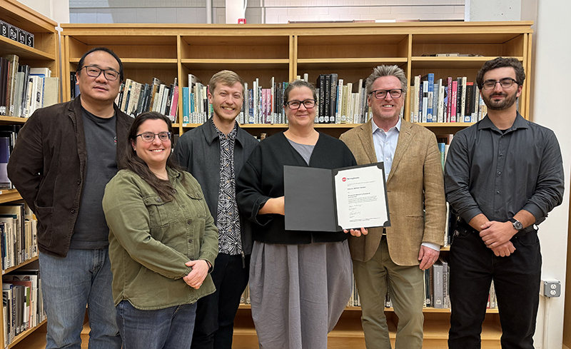 A group of men and women standing in front of a bookshelf with one woman holding an award certificate