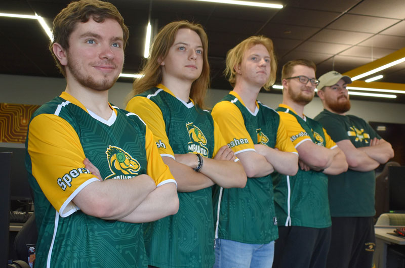 a group of students standing in a diagonal line wearing green and yellow marywood esports jerseys, arms folded, looking off into the distance for a posed photo
